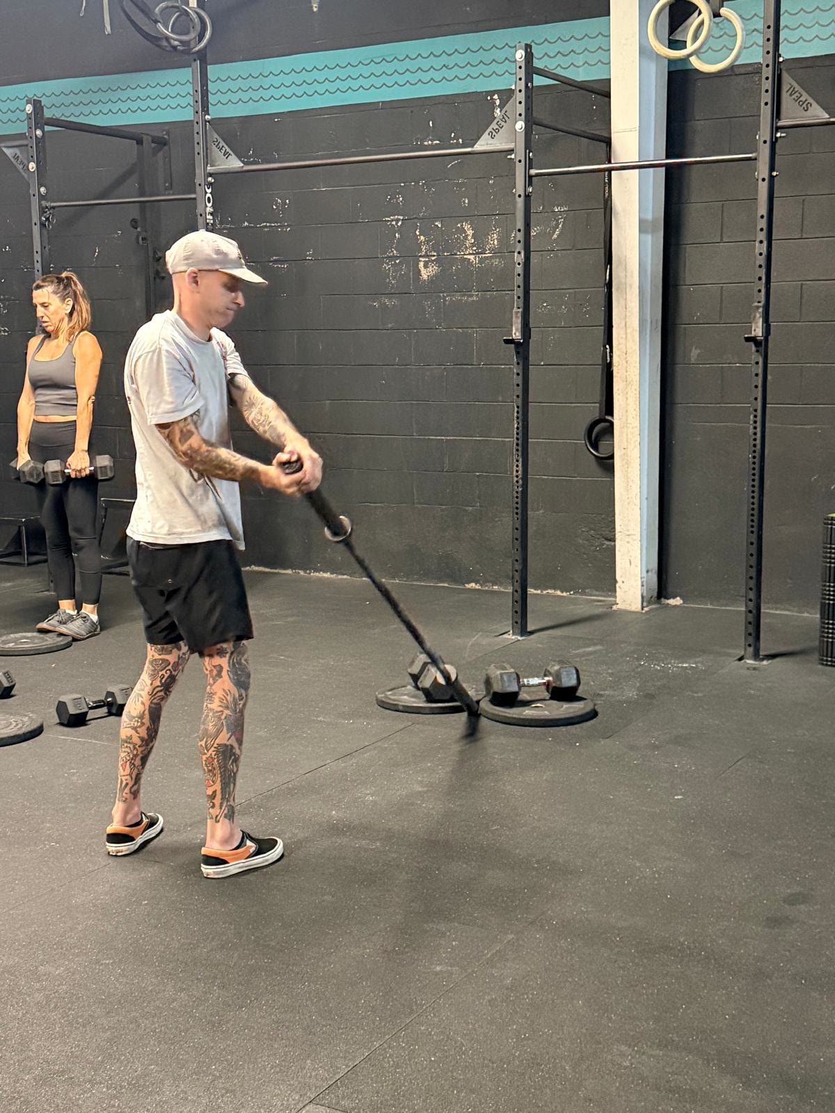 Man doing landmine rotation, woman with dumbbells in background